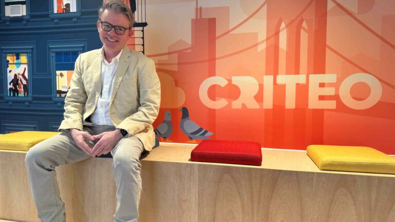A man wearing a light beige blazer and grey trousers sits on a bench with colourful cushions, in front of a bright orange wall featuring the Criteo logo and illustrations of pigeons and a city skyline.