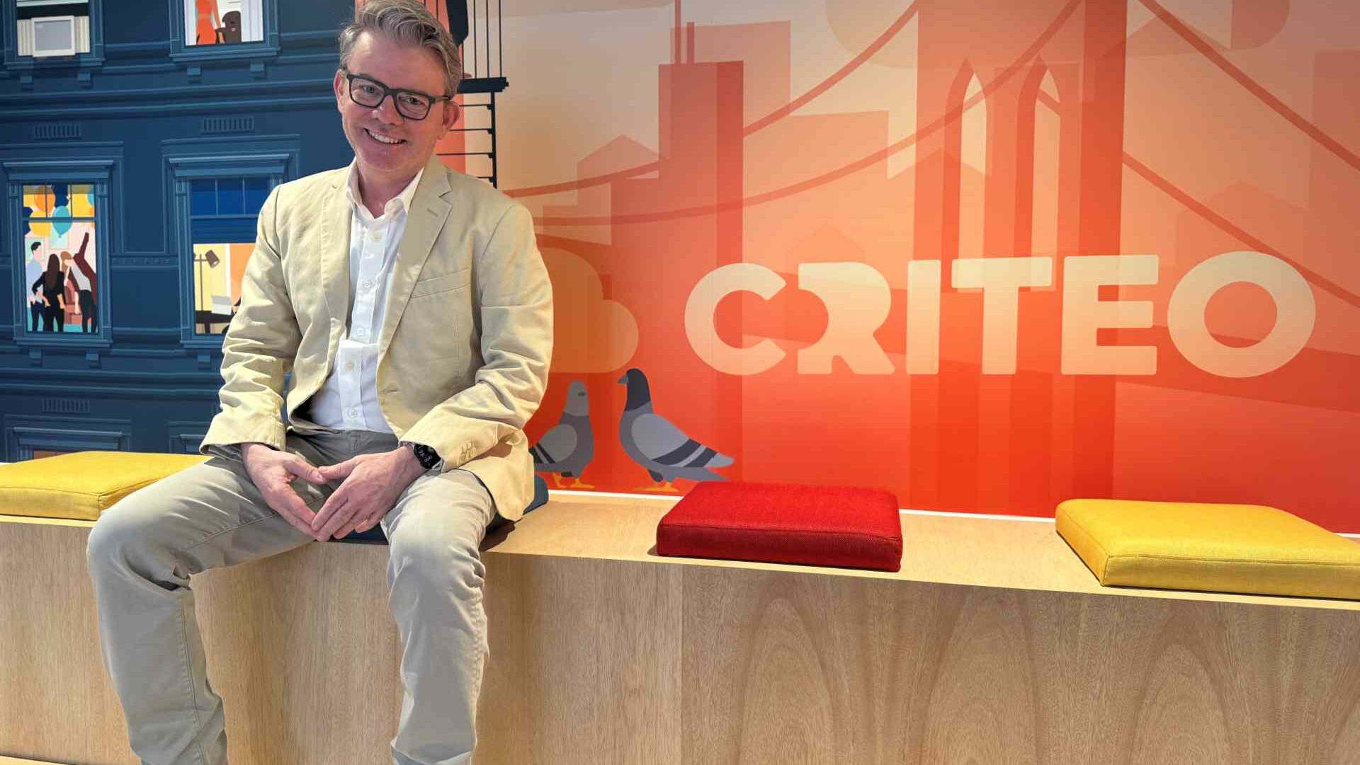 A man wearing a light beige blazer and grey trousers sits on a bench with colourful cushions, in front of a bright orange wall featuring the Criteo logo and illustrations of pigeons and a city skyline.