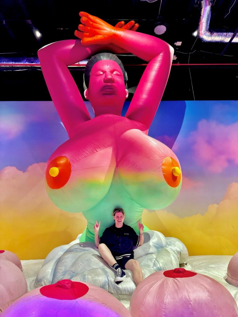 A person sitting in front of a large, colourful inflatable statue of a topless figure with a rainbow background.