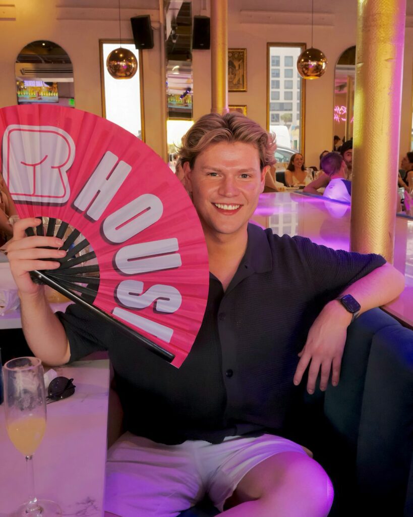 A person sitting in a stylish bar smiling and holding a large pink fan that reads "R HOUSE" with a heart design.