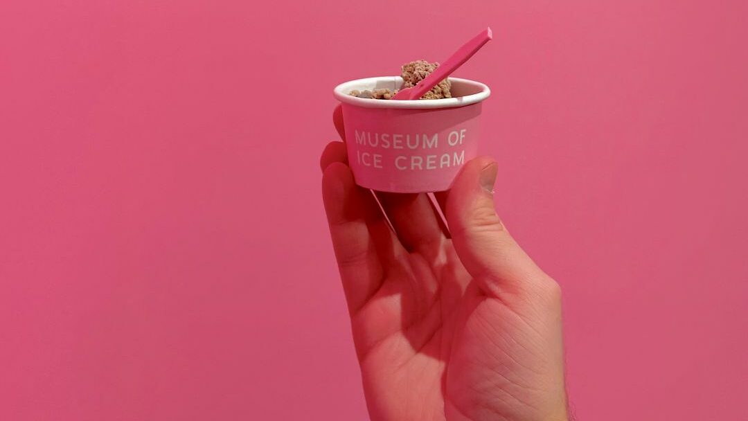 A hand holding a small pink cup of ice cream with a pink spoon, against a pink background. The cup is labelled "Museum of Ice Cream".