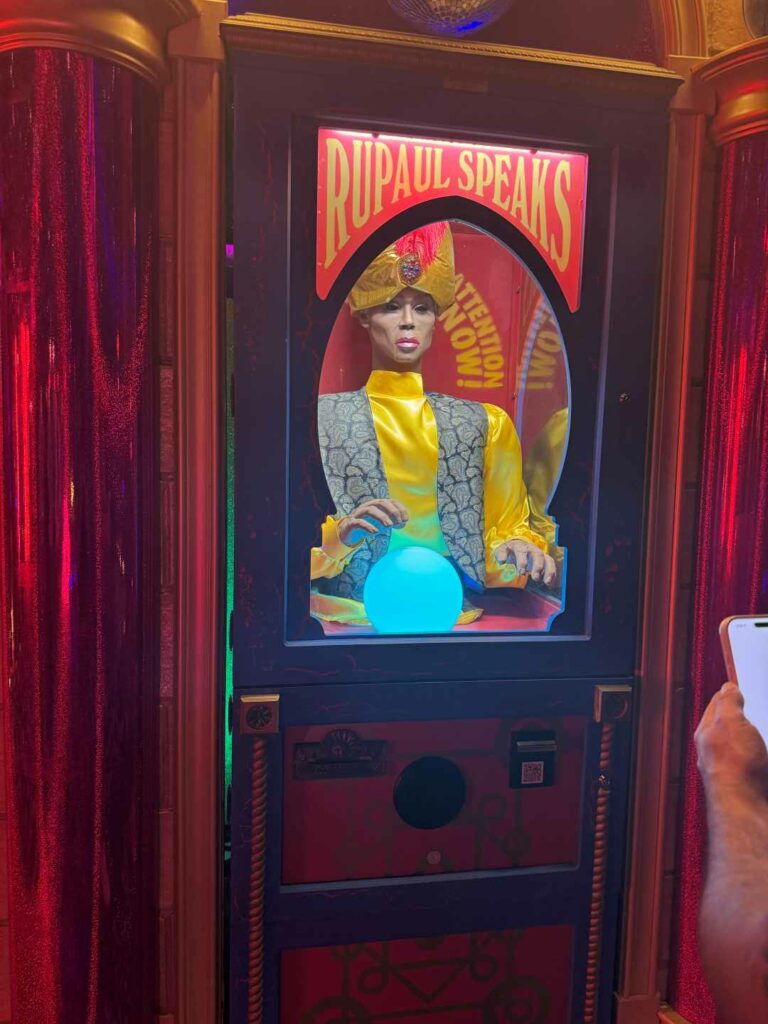 A fortune-telling machine featuring a mannequin of RuPaul dressed in bright clothing, with a glowing crystal ball and the sign "RuPaul Speaks" above.