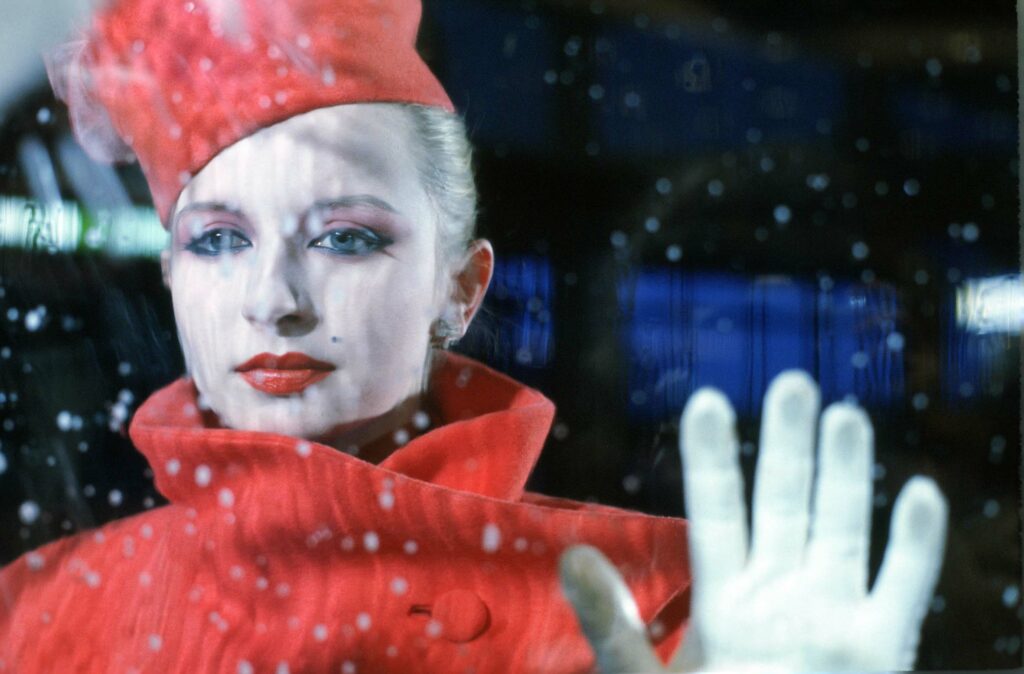 a person in a red coat and hat with white make-up with their hand on a window