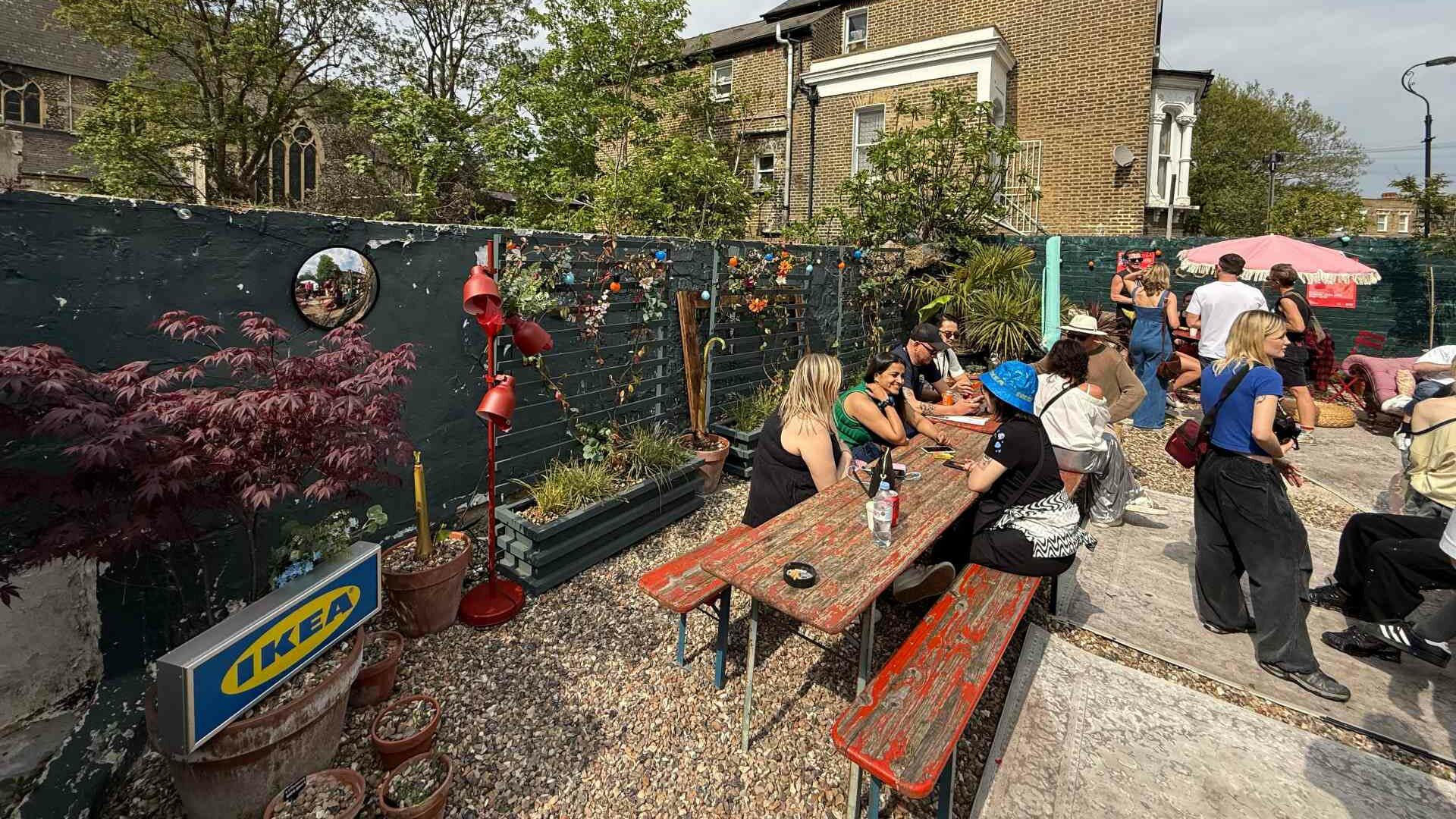People sitting at wooden benches in a sunny outdoor garden area decorated with plants and colourful signs.