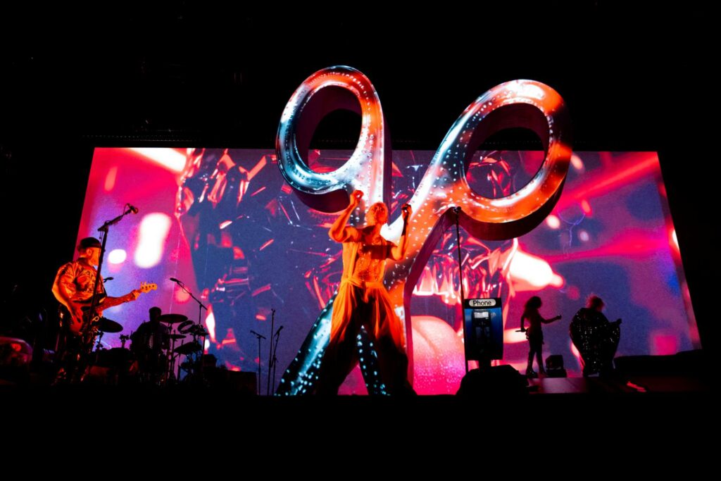 Scissor Sisters performing live in front of a large illuminated scissor sculpture, with vibrant visuals and lighting on a wide screen behind them.