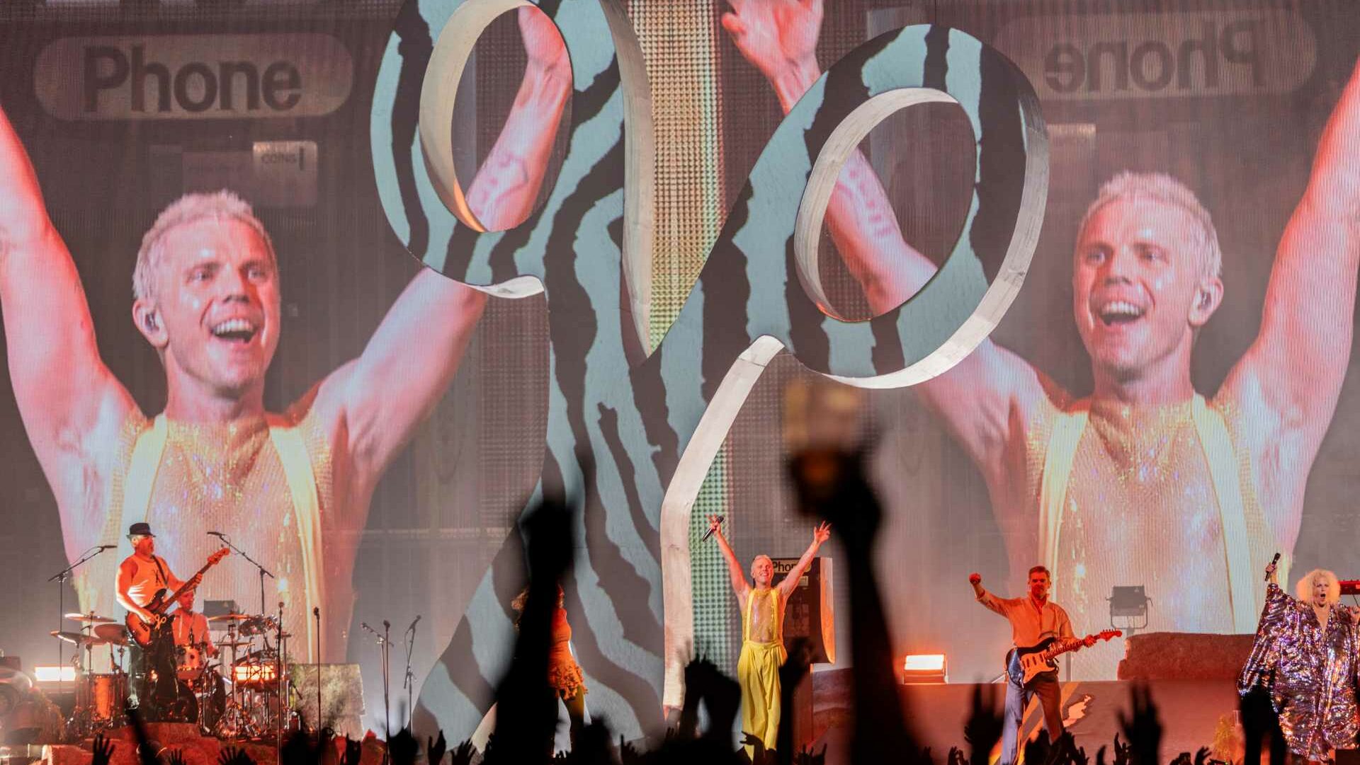 Scissor Sisters performing on stage with a giant scissor logo and projected images of Jake Shears in the background, arms raised, as the crowd cheers.