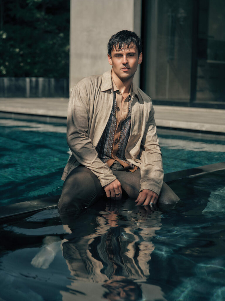 Tom Daley sits on the edge of a pool, wet, in beige Linen overshirt, printed shirt and trousers with leg in pool.