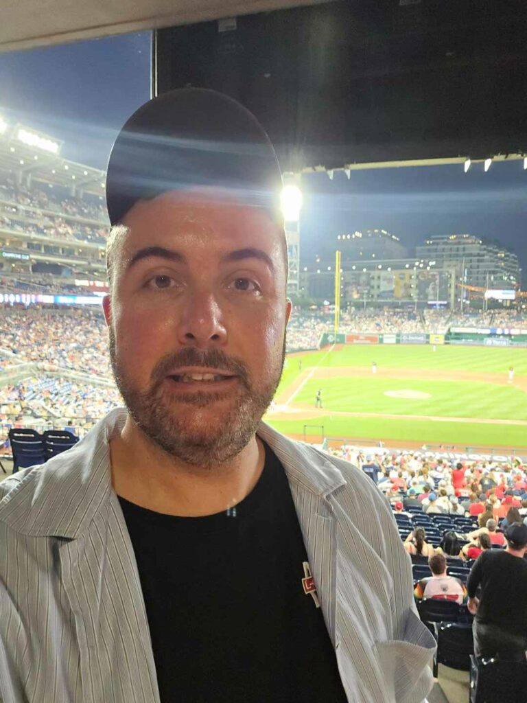 A smiling man looks into the camera with a baseball field behind him