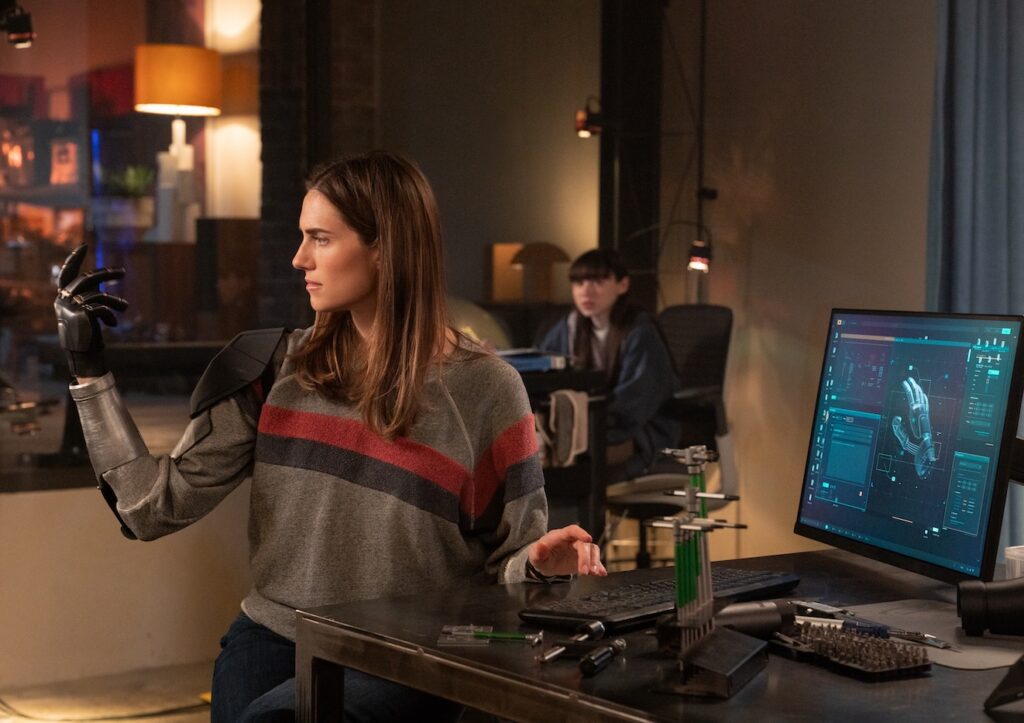Allison William in the film M#GAN 2.0 A woman tests a robotic glove at a workstation, with a 3D model of a hand displayed on a computer screen, while another person looks on in the background.