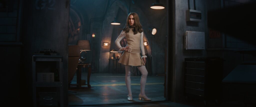 M3GAN stands in a dimly lit, industrial-style room, wearing her signature outfit with hands on hips