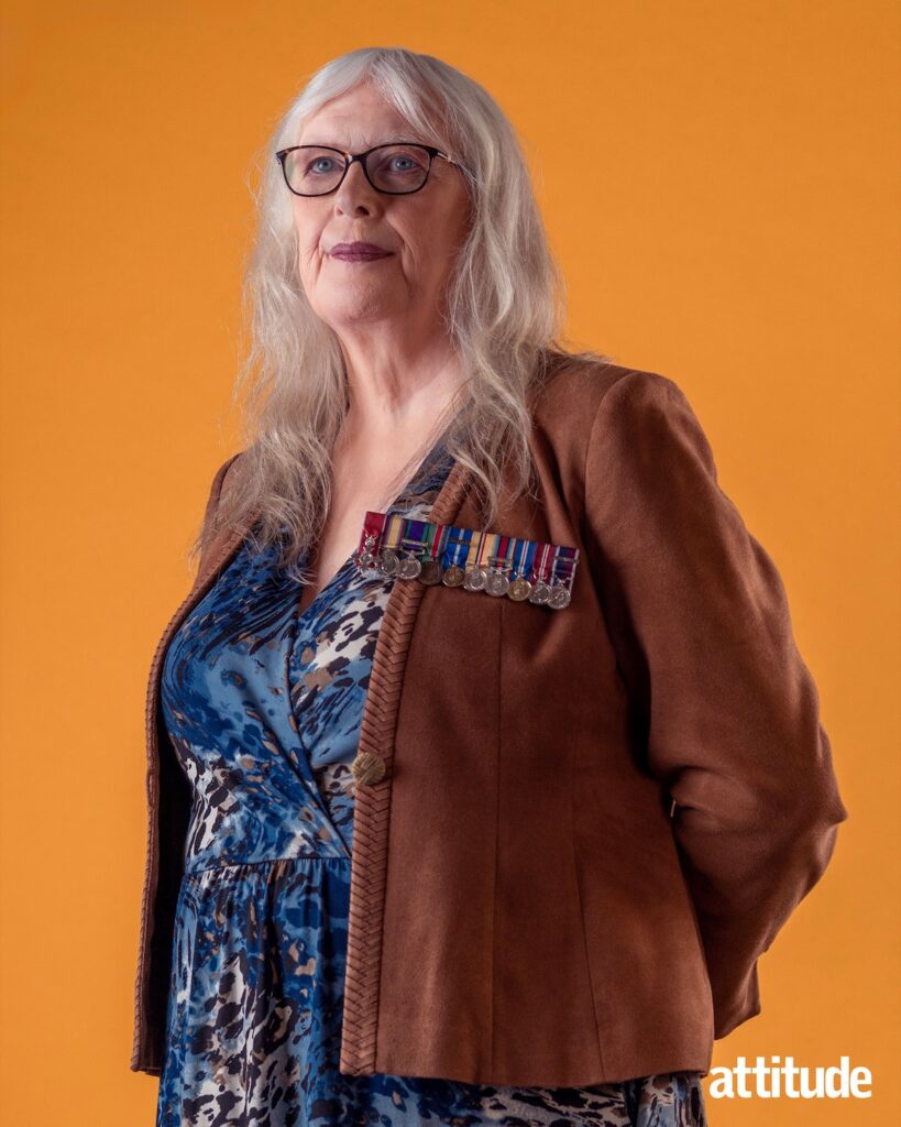 Caroline looking away from the camera, arms behind back, against an orange backdrop wearing a brown jacket and blue top with medals on show and hair down