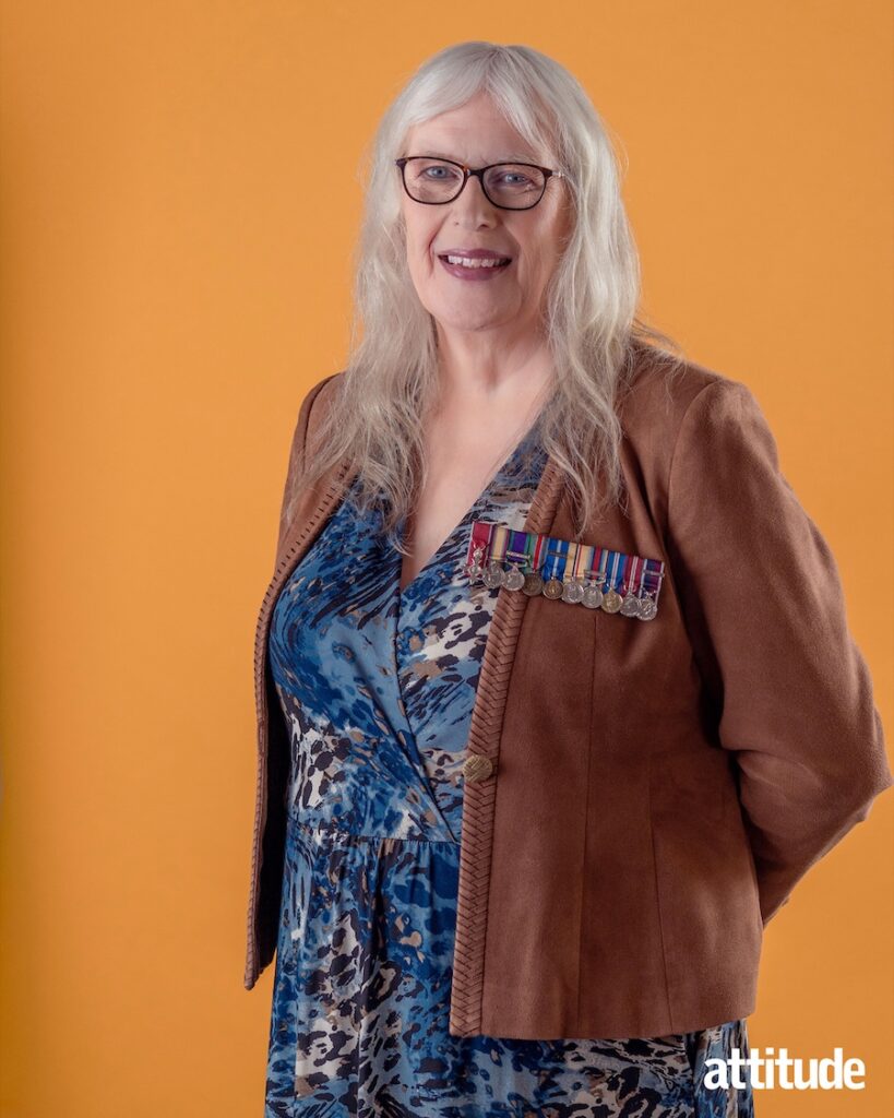 Caroline against an orange backdrop wearing a brown jacket and blue top with medals on show and hair down, smiling