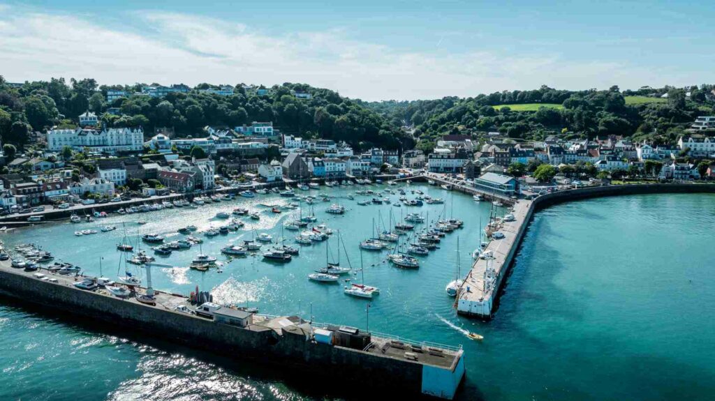 Aerial view of St Aubin’s Harbour in Jersey, with turquoise water, moored boats, and the village nestled against a green hillside in the background.