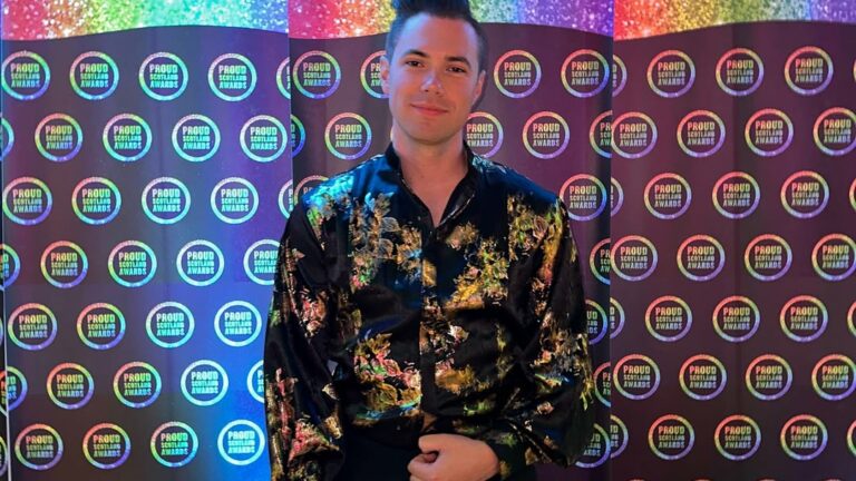 Man standing in front of a rainbow-themed step-and-repeat backdrop, wearing a black floral shirt.