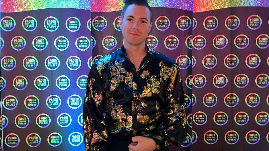 Man standing in front of a rainbow-themed step-and-repeat backdrop, wearing a black floral shirt.