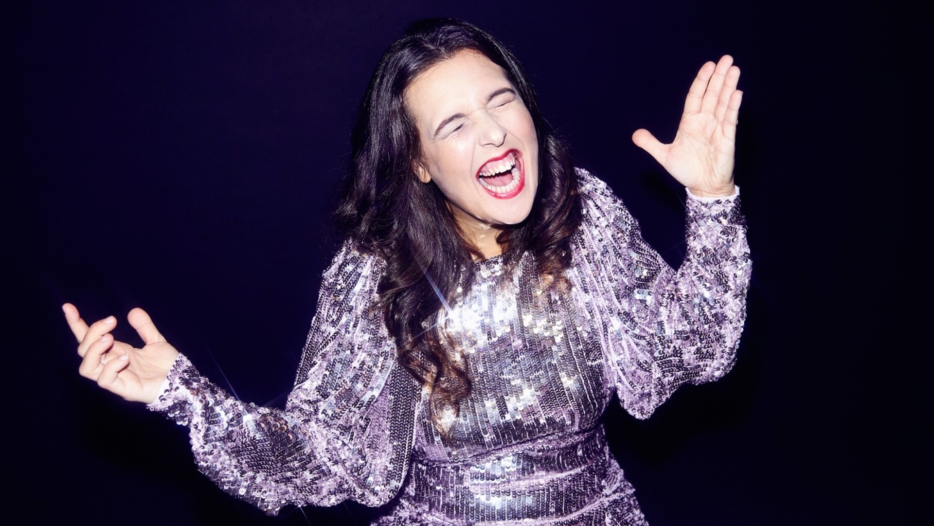 Rosie Jones smiling and gesturing in a sparkly outfit against a dark background