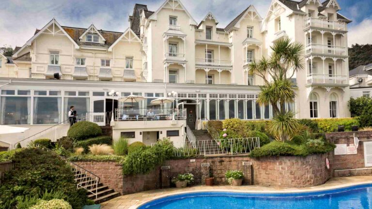 The exterior of the Somerville Hotel in Jersey, a grand white Victorian building with gabled roofs, large windows, and a terraced garden overlooking an outdoor pool