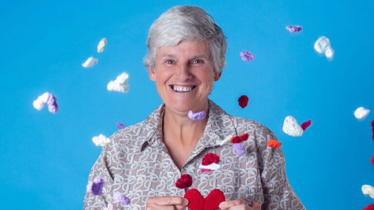 Head and shoulders of Tracey Howe against a blue backdrop, with knitted multi-coloured hearts being thrown in the air