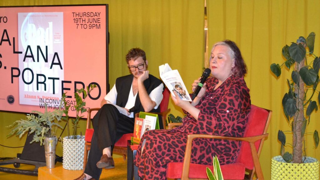 Alana S. Portero and Jorge Garriz sat down on red chairs at Second Home, Alana reading her book Bad Habit aloud to the audience at the Romancero Books Fifth Anniversary event