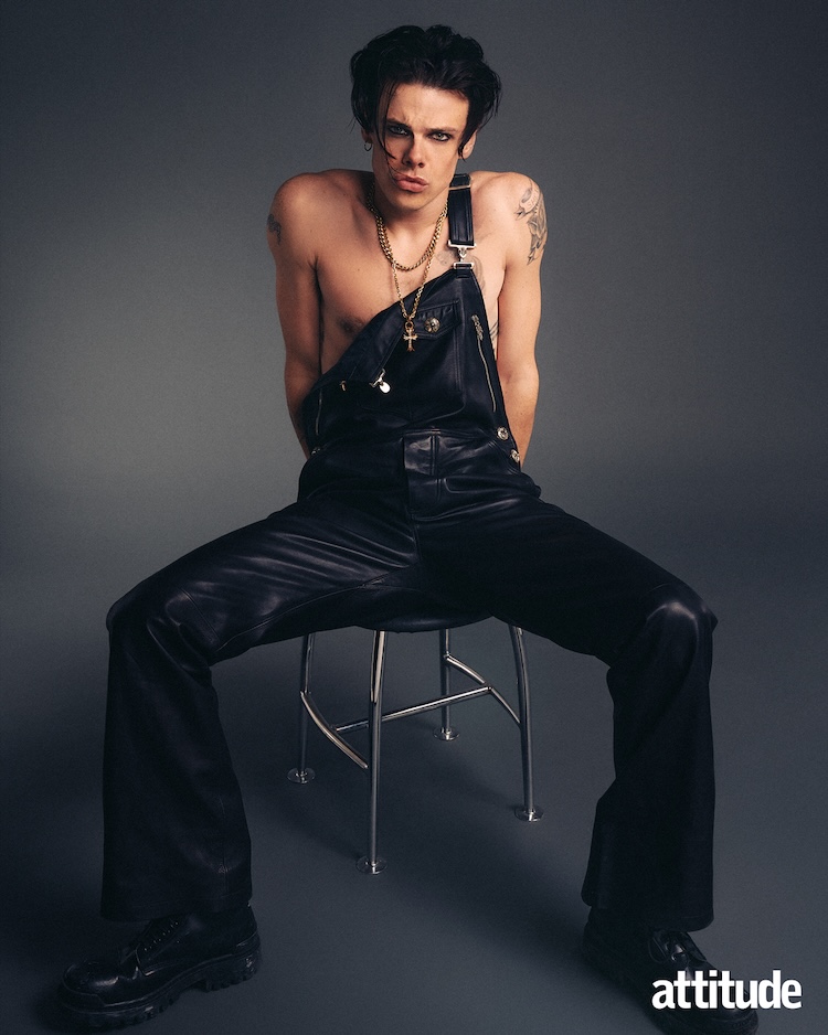 Yungblud sitting in a pair of dungarees with one strap undone, grey background