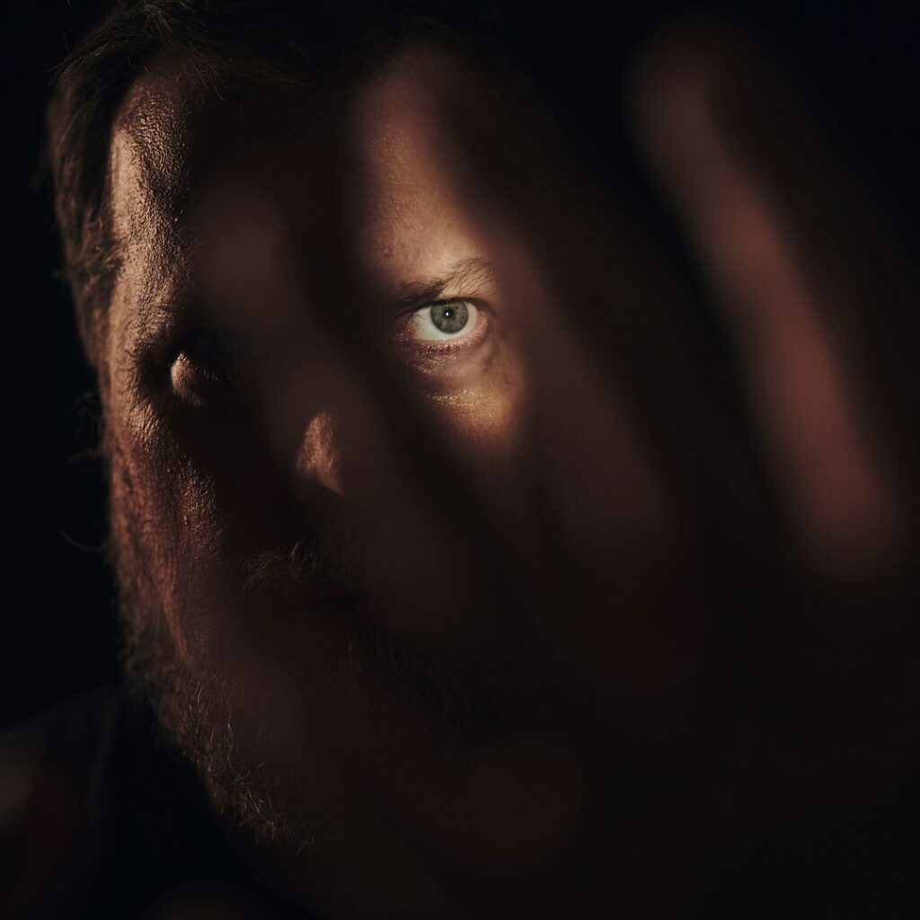 Close-up of John Grant’s face, partially obscured by a hand, with one eye dramatically lit in shadow