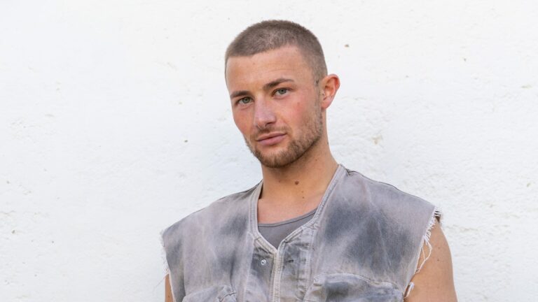Press shot of Jordon Burrow wearing a grey sleeveless top looking at the camera smirking infant of a white wall