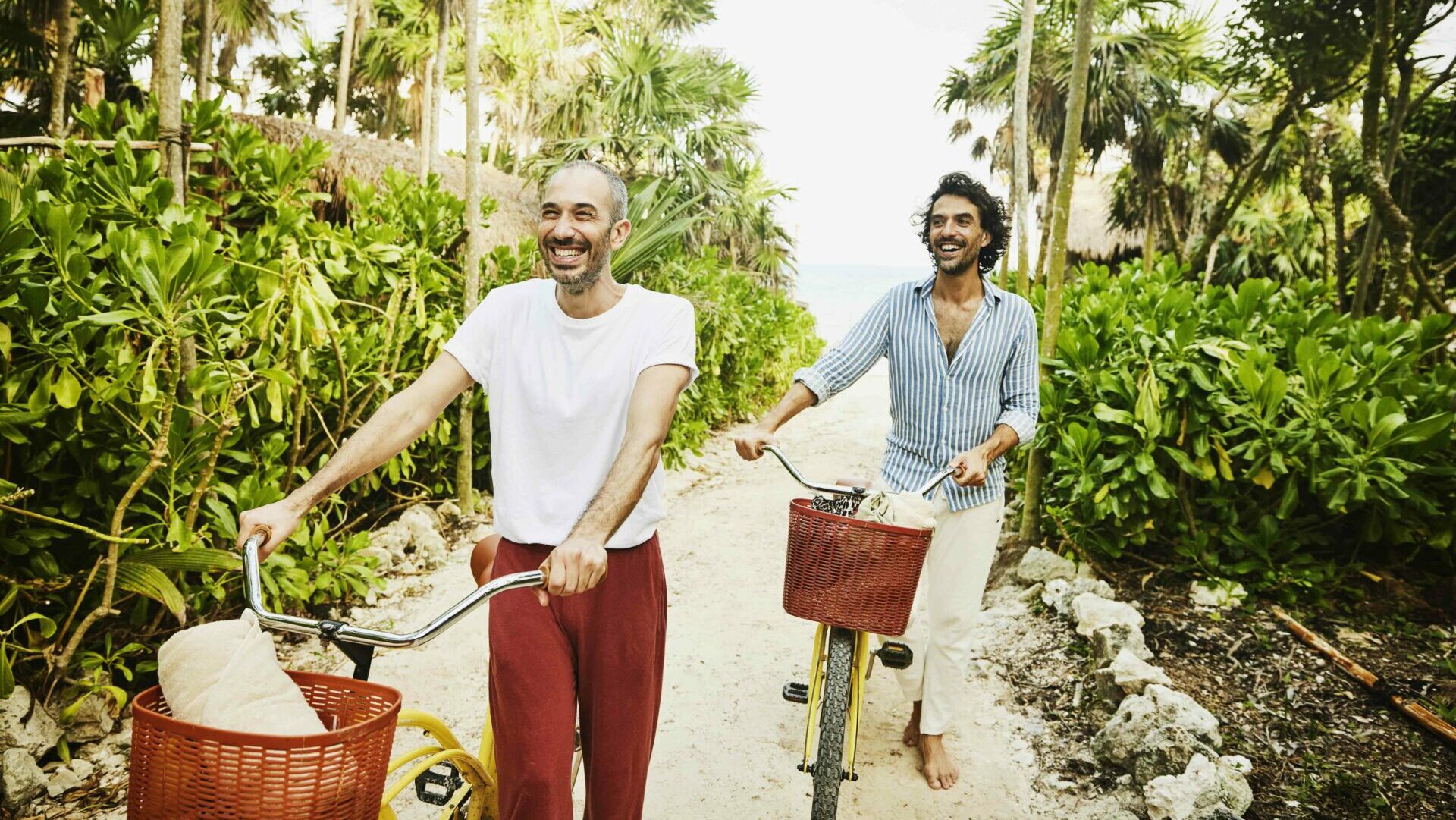 Medium wide shot of laughing gay couple walking bikes on path from beach at tropical resort