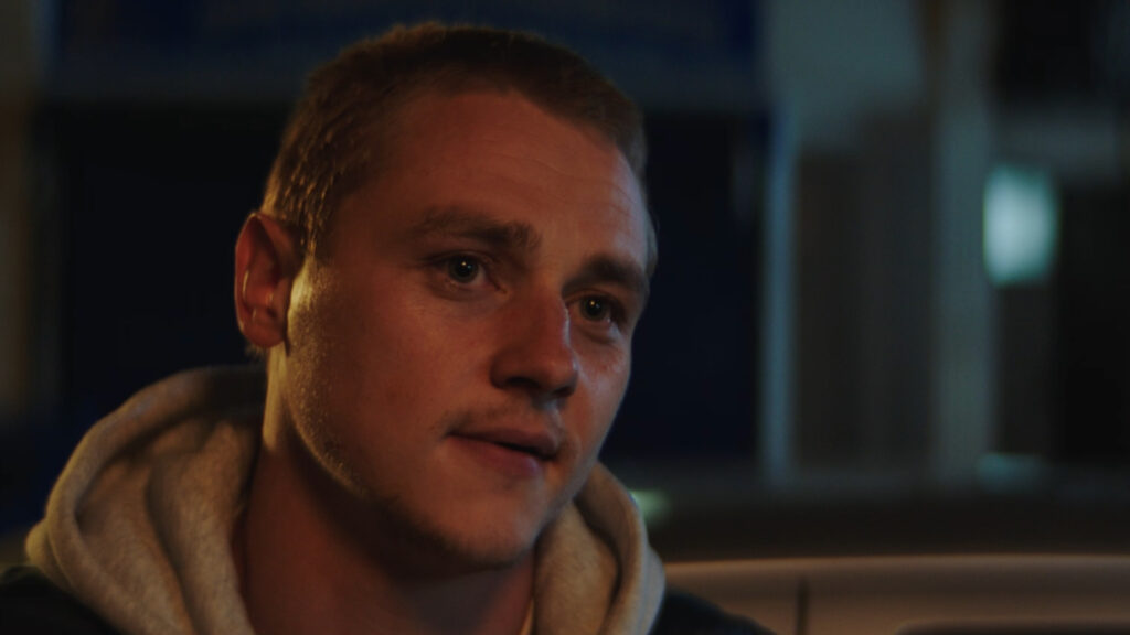 Close-up of a young man with a buzz cut wearing a hoodie, sitting outside at night. His expression is thoughtful and slightly emotional, lit by soft, warm lighting.