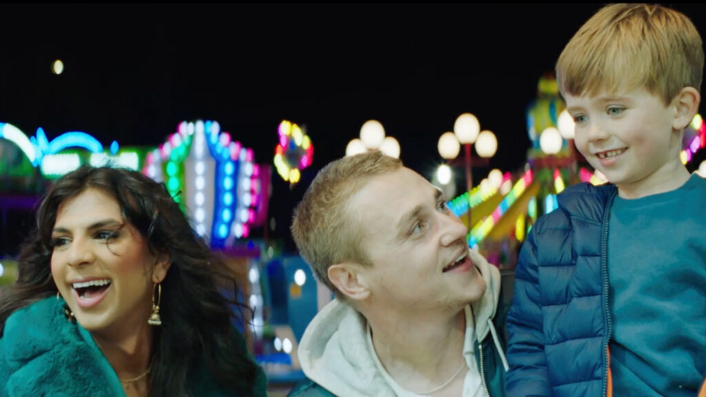 A trio at a vibrant fairground at night: one wearing a teal coat, a smiling man in a hoodie, and a laughing young boy in a puffer jacket. 