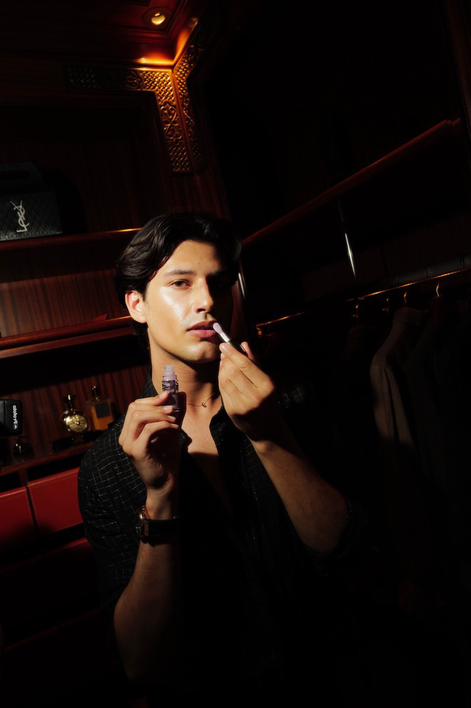 Zak Heath applying lip gloss, he is it by the flash of the camera in front of a rich dark back-drop