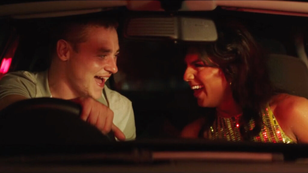 Inside a car at night, two people laugh together under red lighting. One wears a sequin gold outfit, while the other grips the steering wheel, leaning toward their companion with a wide smile.