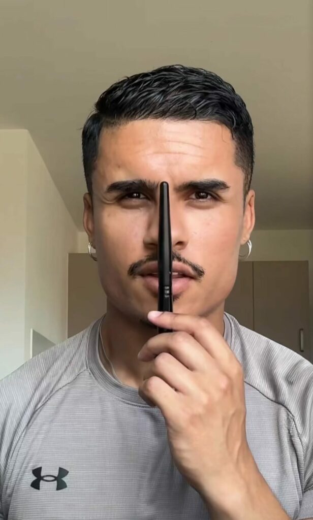 Syed Ahmed wearing a grey t-shirt, with half of his face painted with subtle makeup, the other half natural, holding up a makeup brush as a divider