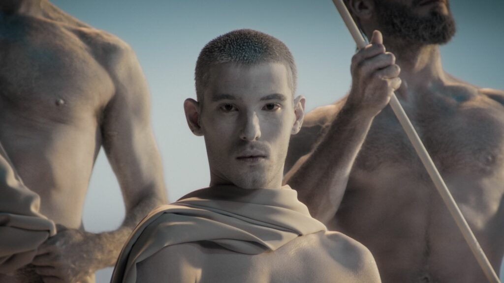 Person painted like a statue, wearing a toga and looking directly at the camera, with other statue-like figures behind