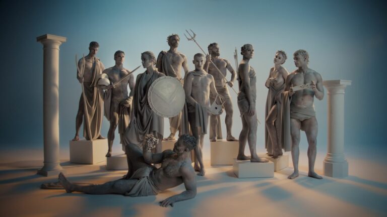 A group of people painted like stone statues, dressed in togas and posing with props like shields and grapes, surrounded by pillars in a studio setting.