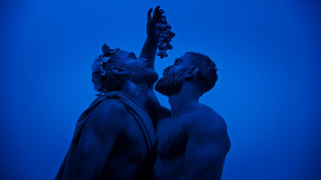 Two men dressed as Greek gods embrace while holding a bunch of grapes over their heads
