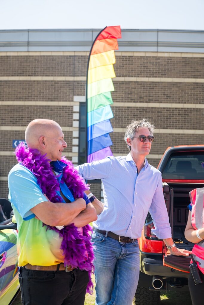 Two men, one dressed smartly in a blue shirt and sunglasses the other wearing a rainbow t-shirt and a purple feather bower 