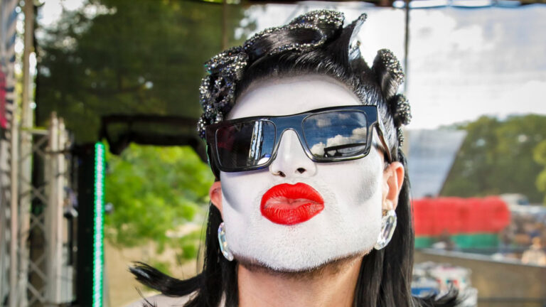 Danny Beard in drag wearing sunglasses blowing a kiss to camera wearing red lipstick