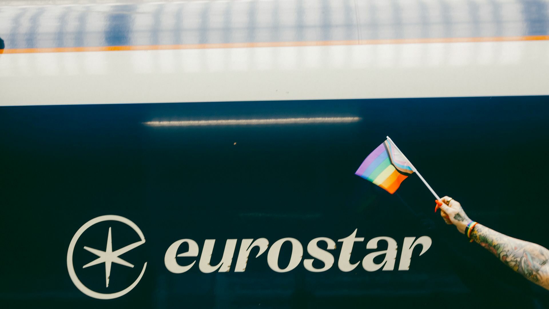 A hand holding a Progress Pride flag in front of a Eurostar train.