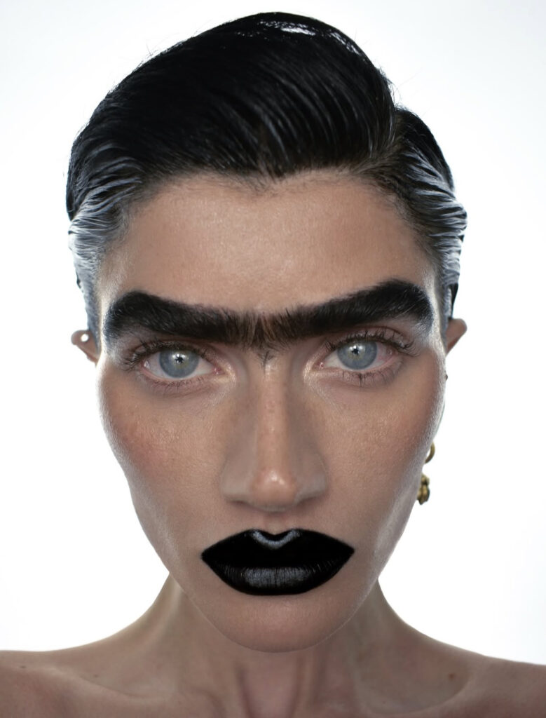 Sophia Hadjipanteli with slick back black hair, with a bushy uni-brow and bold black lipstick