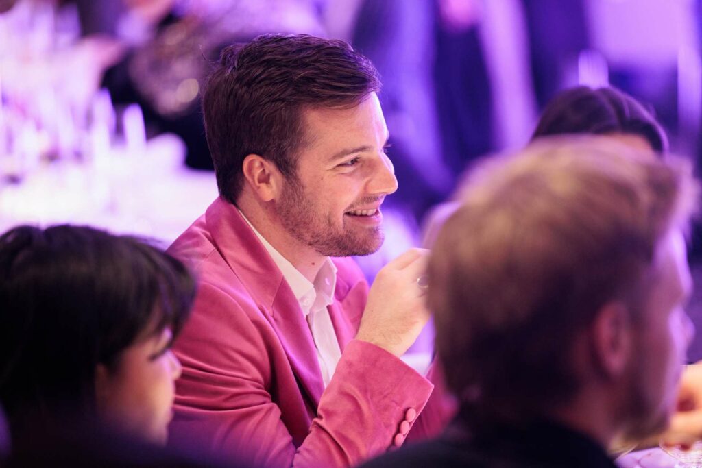 A smiling man in a pink business suit sitting at a table at an event