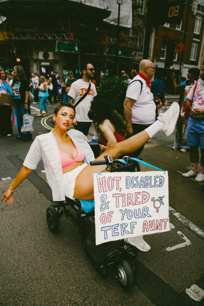 a person in an open white shirt, shorts and pink bralet on a mobility scooter with the sign 'Hot, disabled and tired of your terf aunt.'