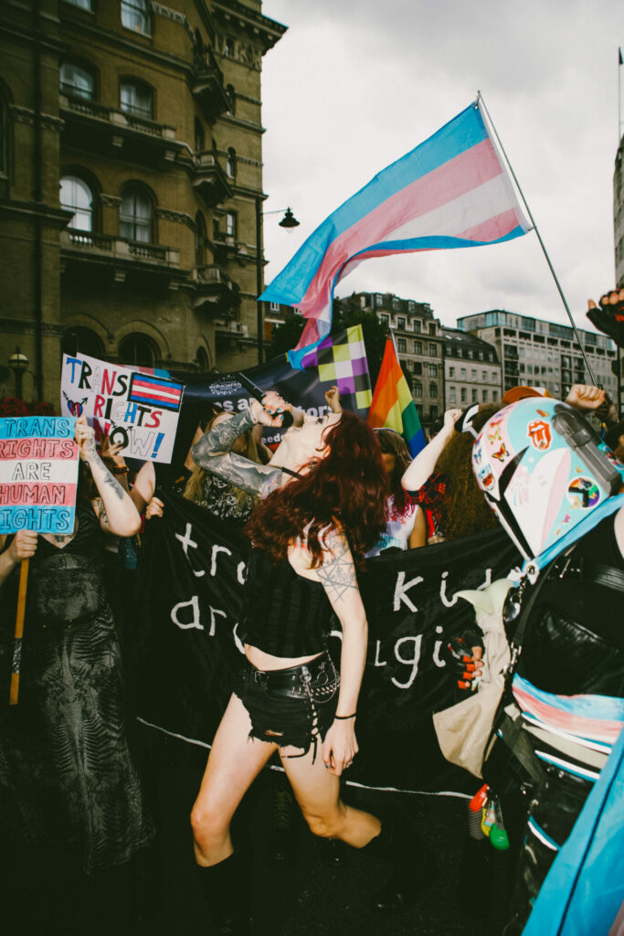 a tattooed person in a black outfit leading chants into a megaphone. Behind them, a group holds a black banner reading "trans kids are magic" and waves transgender and rainbow flags.