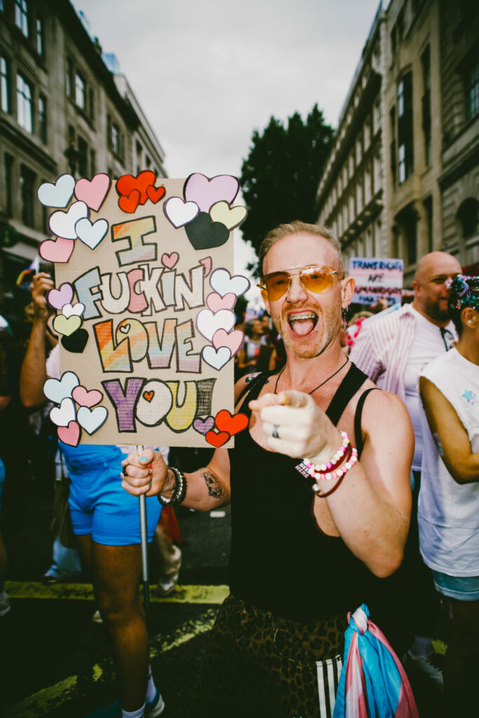 A person wearing a black vest holding a placard saying “I fucking love you” with people behind him