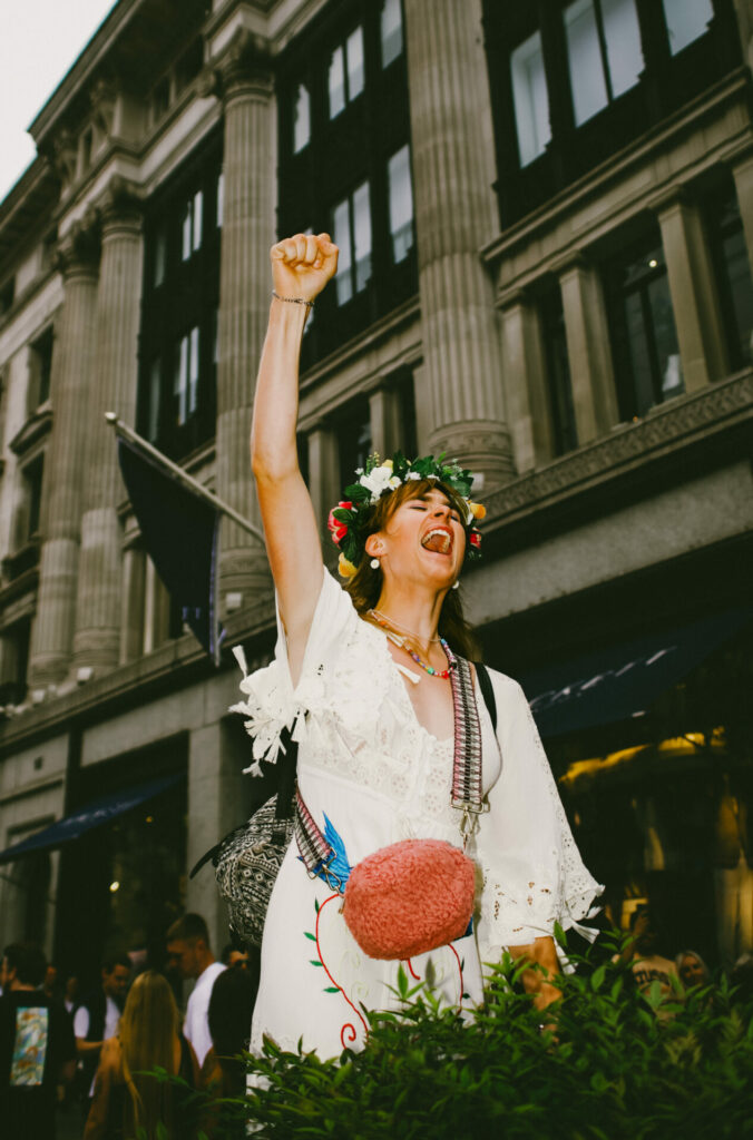 Someone at trans pride stood tall with their first punching the air protesting for trans rights. They wear a flower crown and a white dress.