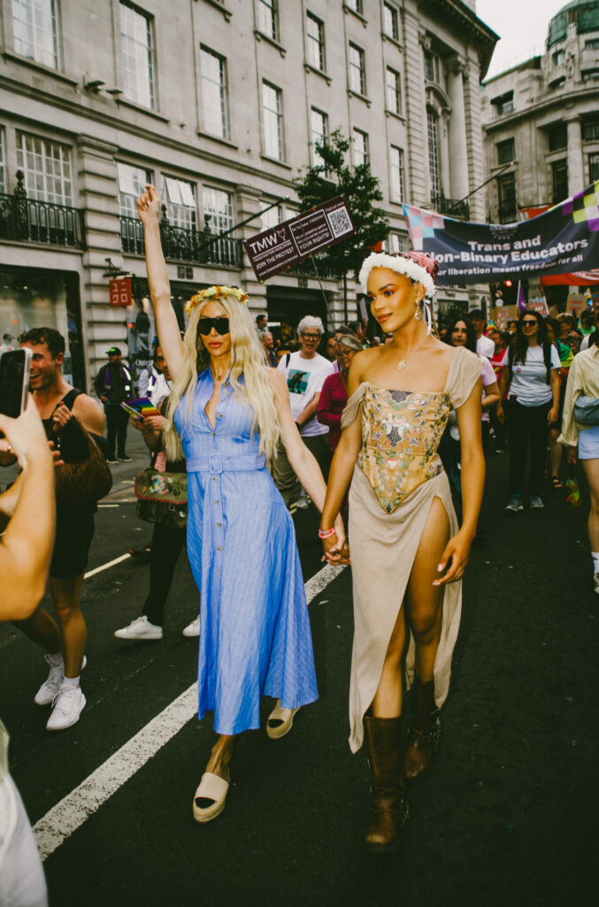 Two trans women walk hand in hand at a Trans Pride parade, smiling and radiating confidence. One wears a long blue dress and sunglasses with a flower crown, while the other wears a floral corset dress with a thigh-high slit and boots.