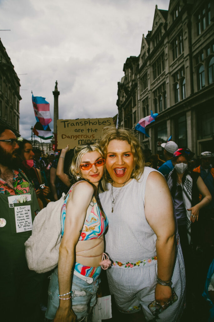 Two trans individuals pose joyfully for the camera at a Trans Pride march. One wears pink sunglasses and a colorful crop top with denim shorts, while the other wears a light romper with a floral belt.