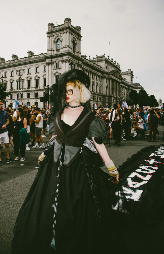 A protester wearing a black gown with a train stretching the streets of London with the words 'Trans + Here' written.