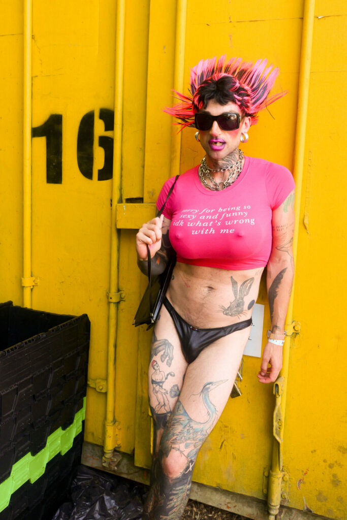 Man in a thong and a cropped pink top wearing a breast plate and sunglasses leaning up against a yellow backdrop