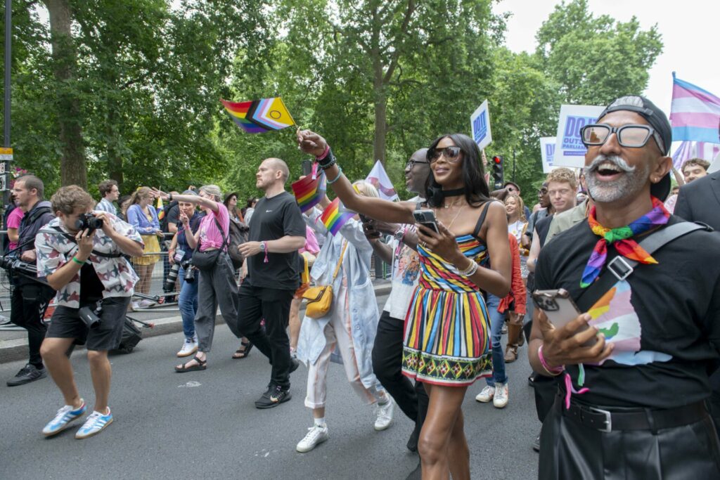 Naomi Campbell in a multicoloured striped dress waving a Pride Progress flag with other marchers