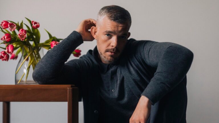 Russell Tovey in grey top with flowers behind him (Image: Rolling Stone UK)